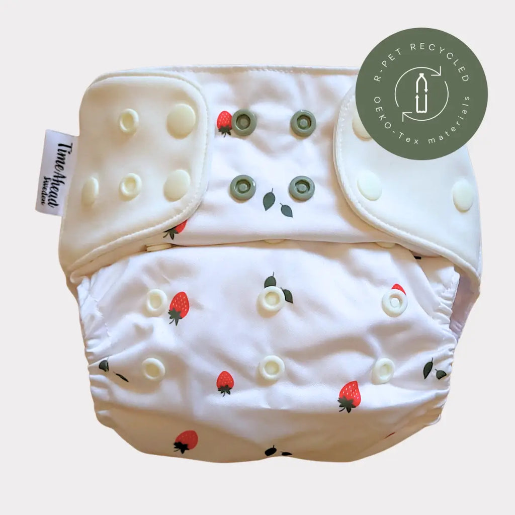 Vit återanvändbar tygblöja med jordgubbs- och bladtryck, med tryckknappar i krämigt och olivgrönt.