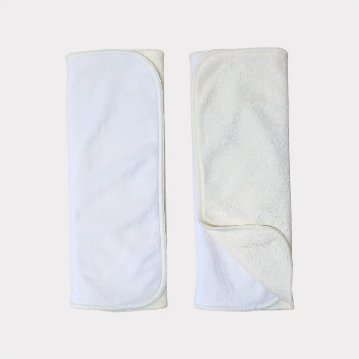 Två vita, mjukt texturerade tygblöjinlägg med en subtil ribbad kant och prydligt vikta hörn.