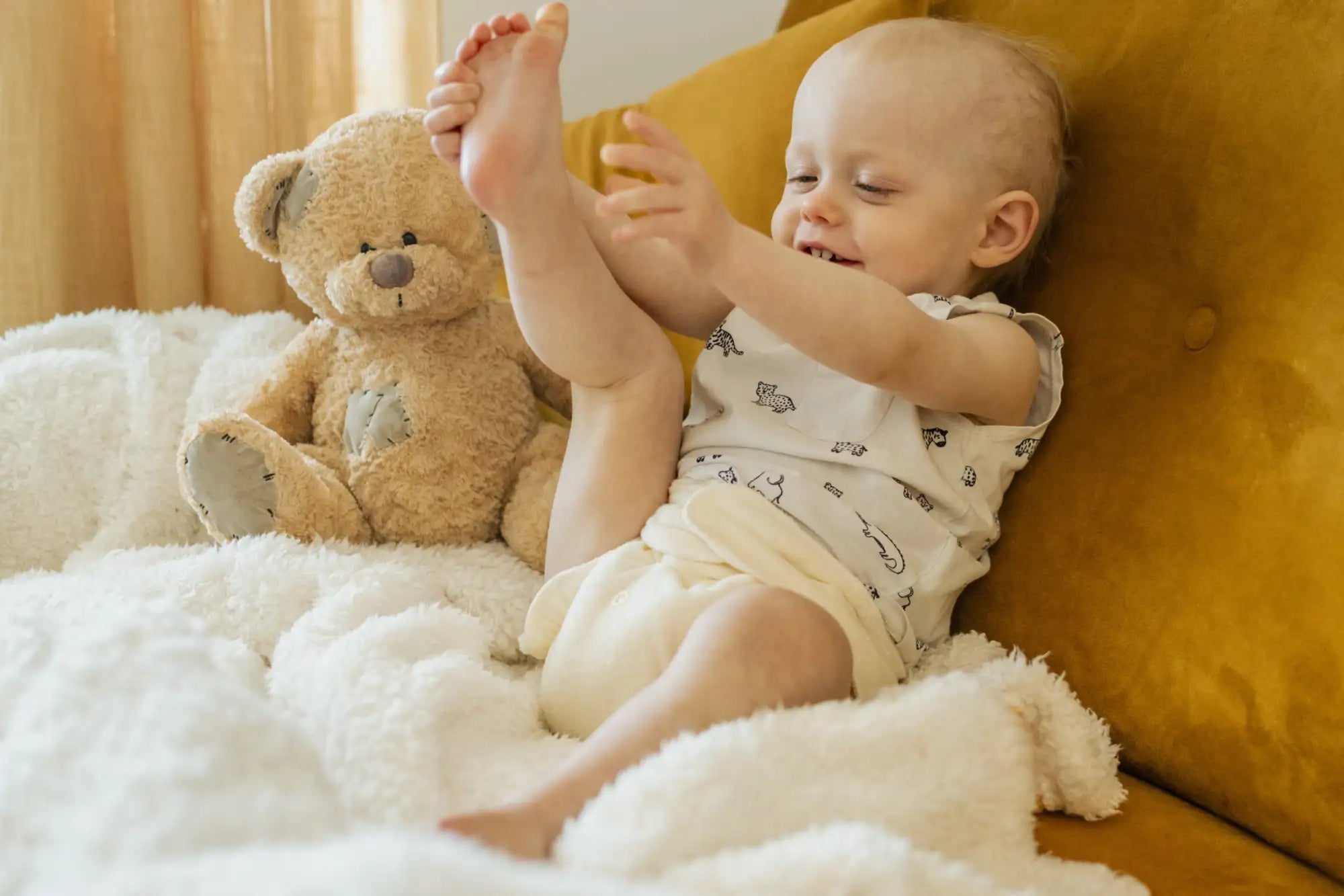 En mjuk, ljusbrun teddybjörn med en broderad näsa och lapptäcksdetaljer sitter bredvid en leende bebis på ett fluffigt vitt täcke.