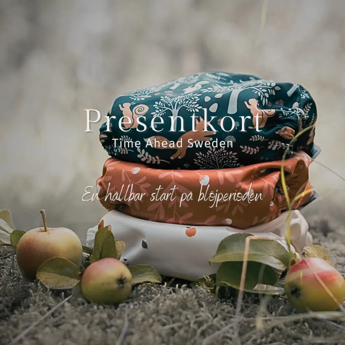 Stapel med tre hopvikta tygblöjor, med ett mörk turkos mönster med skogsdjur och träd på toppen, ett orange blommönster i mitten och en vit bas med små röda äpplemotiv längst ner.