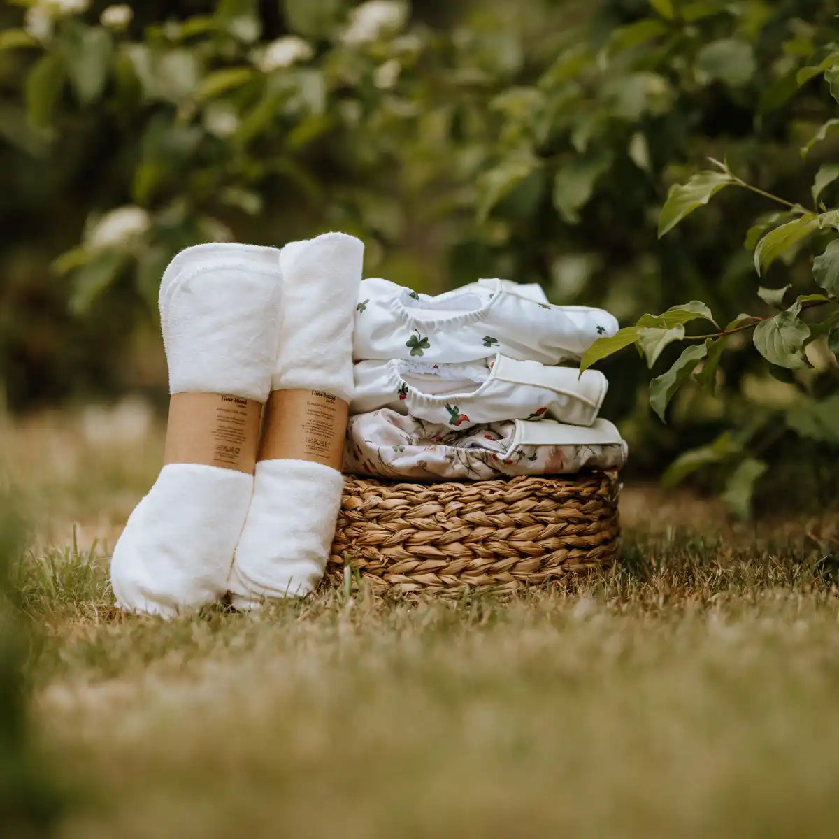 Stapelfynd med vita, rullade tygartiklar med bruna etiketter bredvid en liten, vävt korg som innehåller vikta babykläder.